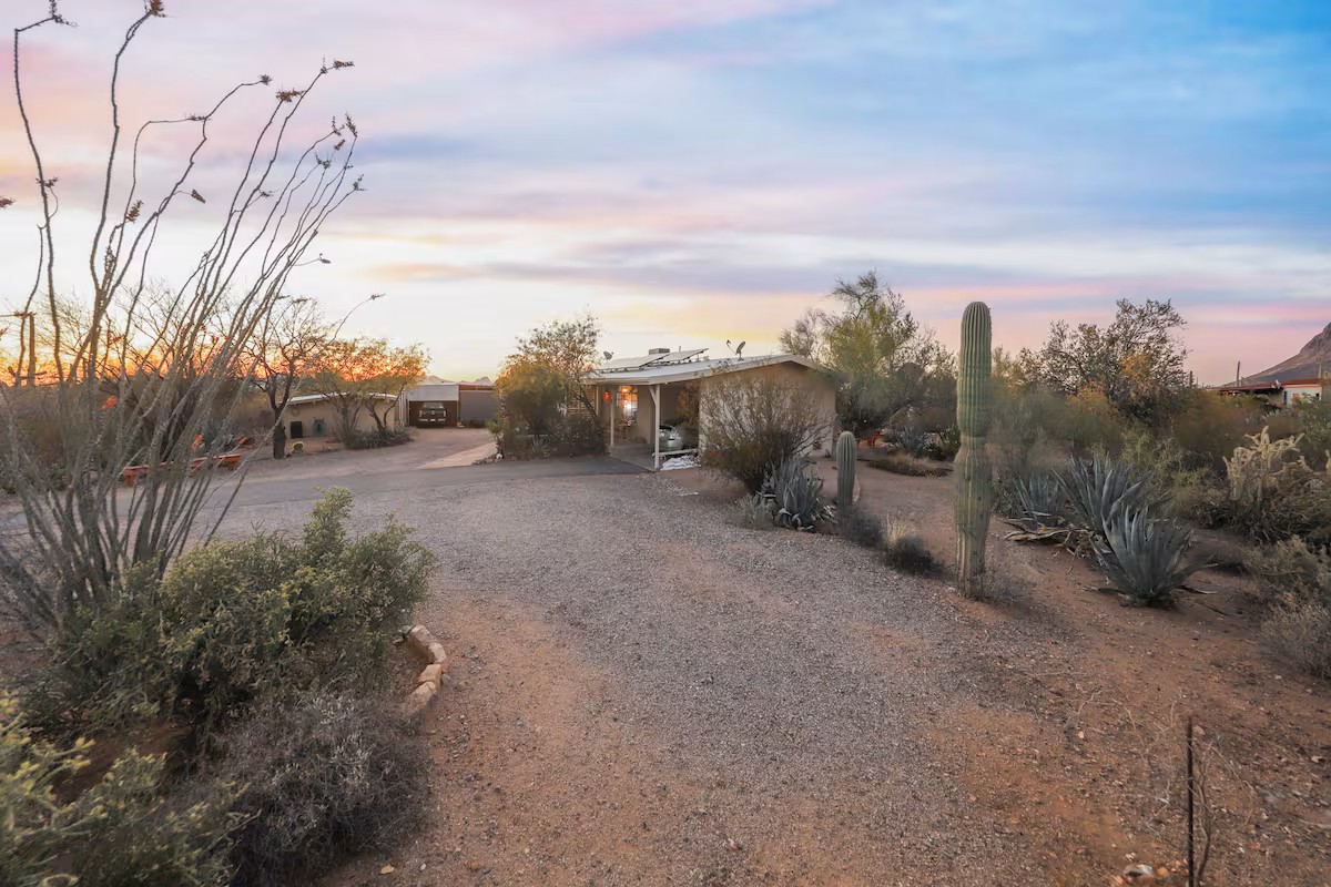 Desert landscape at sunset featuring a saguaro cactus, gravel pathways, native plants, and a small casita in the background.