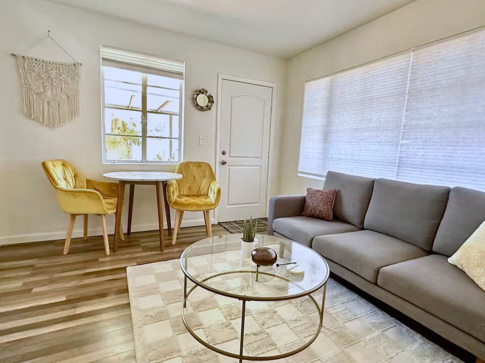 Sunny guesthouse living area with light wood floors, a gray sofa, yellow accent chairs, and natural light streaming through large windows.