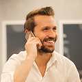 Smiling man with beard and white shirt talking on a smartphone indoors.
