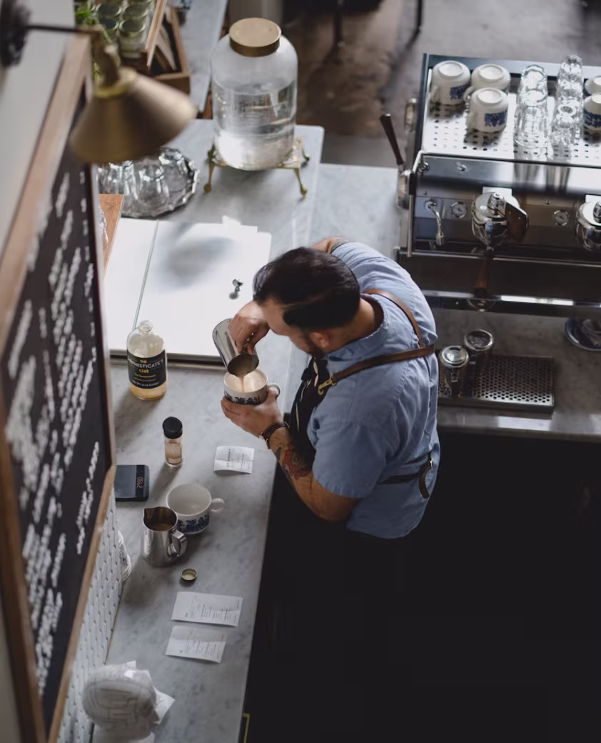 Barista wearing a blue shirt and apron pouring milk into a cup behind a coffee counter.