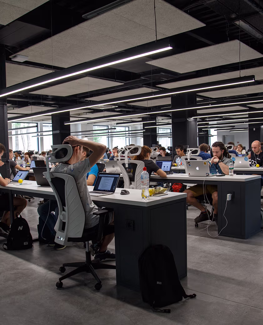 Modern open office space with multiple people working at desks on laptops under linear ceiling lights.