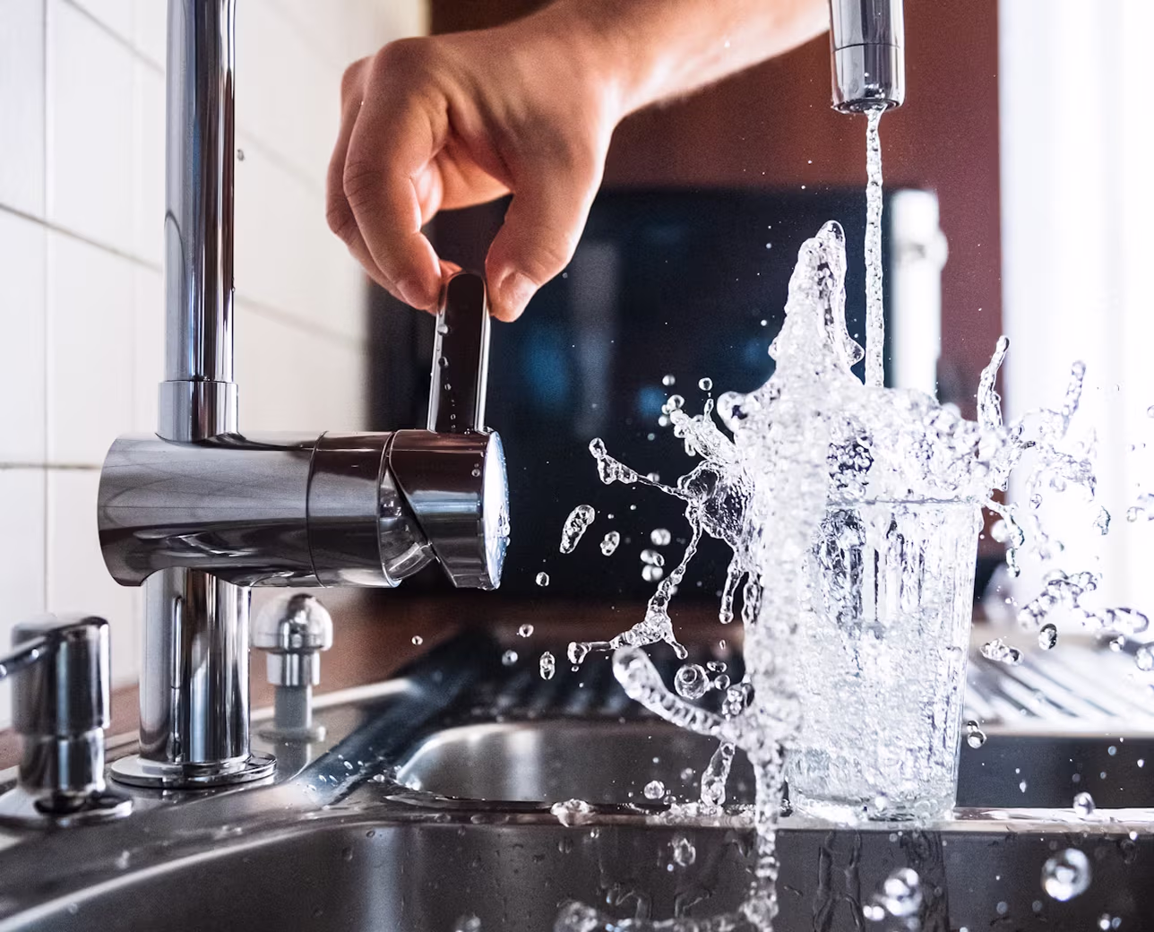 Hand turning off a kitchen tap with water splashing out of a glass under the running faucet.