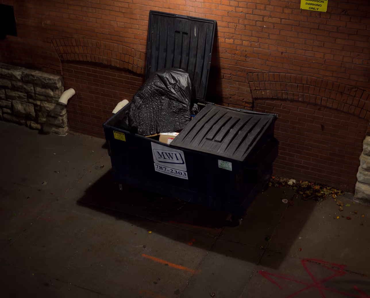 Open black dumpster filled with trash bags and boxes next to a brick wall on a street at night.