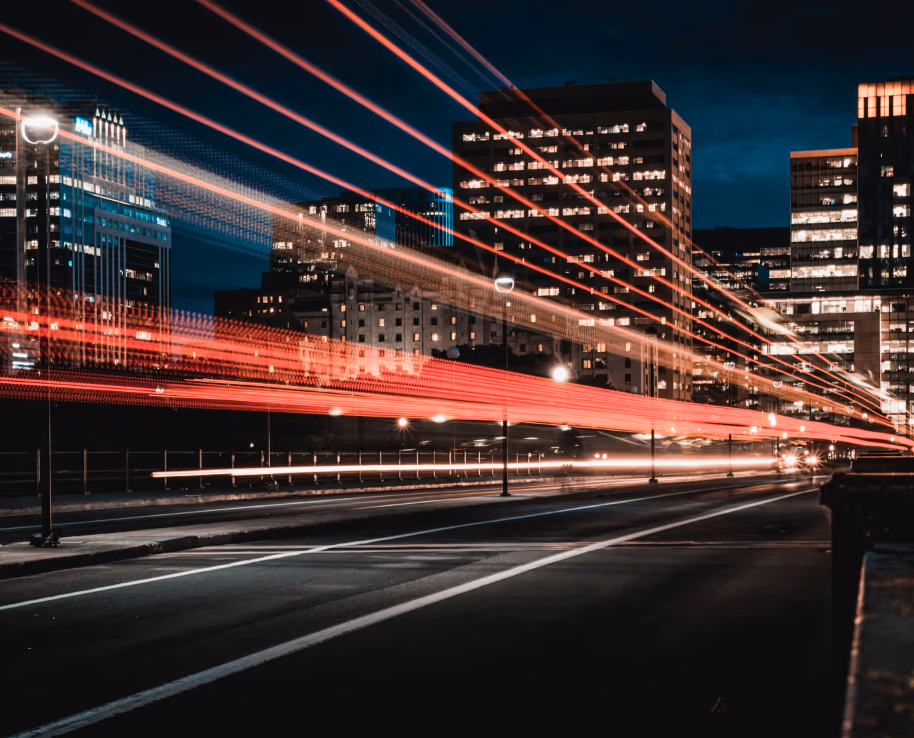 Long exposure night shot of a city street showing red and white light trails from moving vehicles and illuminated office buildings in the background.