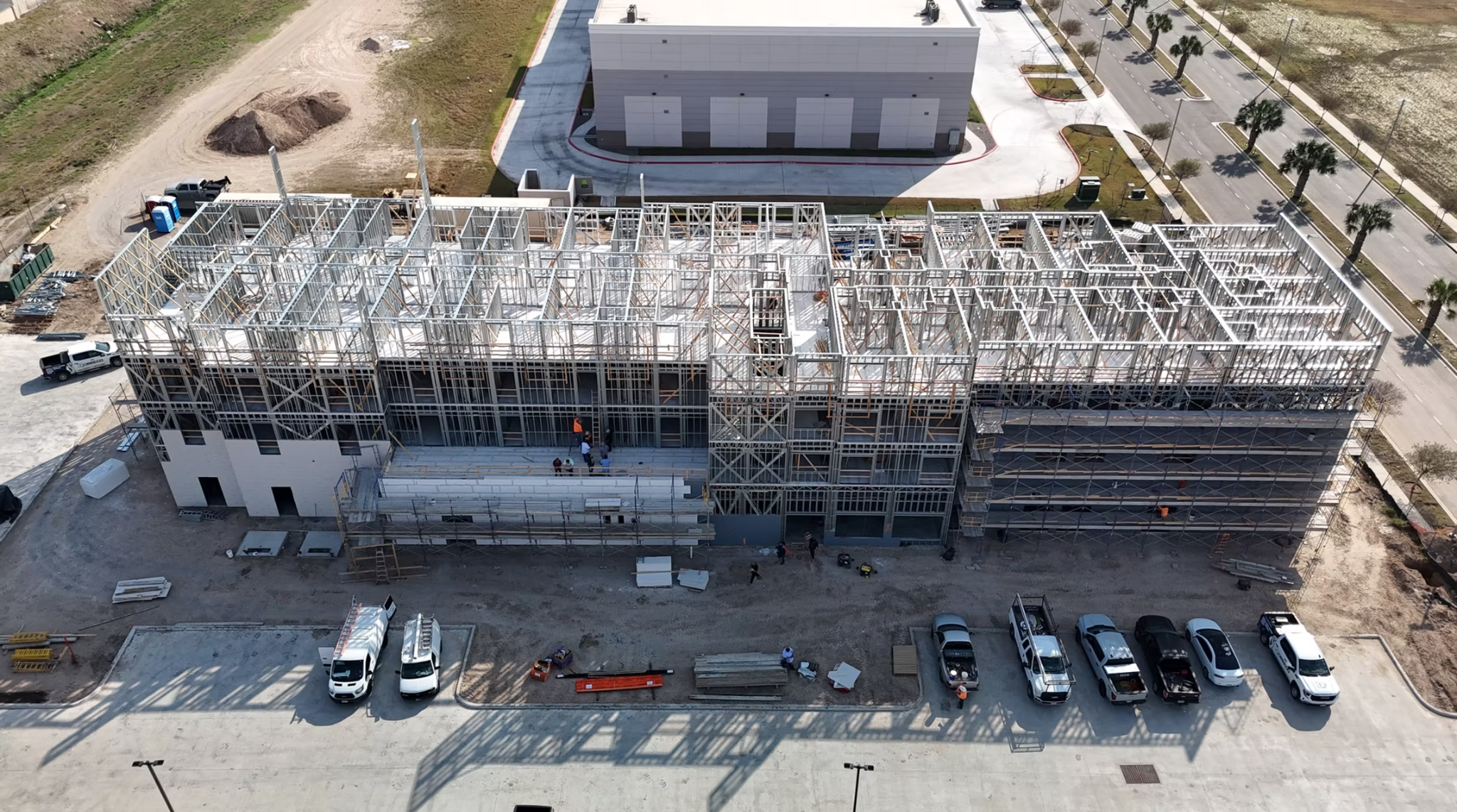 Aerial view of a mid-rise building under construction with light gauge steel framing installed across the upper floors, scaffolding along the right elevation, and construction vehicles and materials staged below on a paved lot.