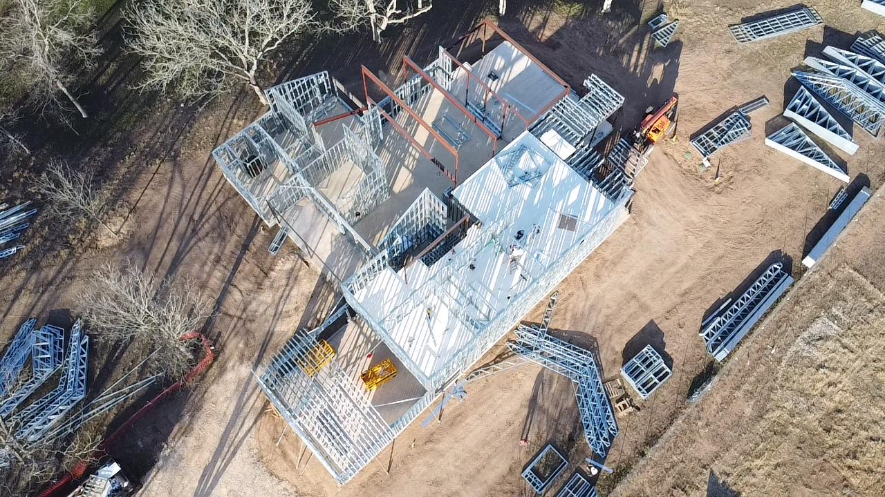 Aerial view of a custom home under construction with cold-formed steel wall panels, roof trusses, and staged framing components on site.
