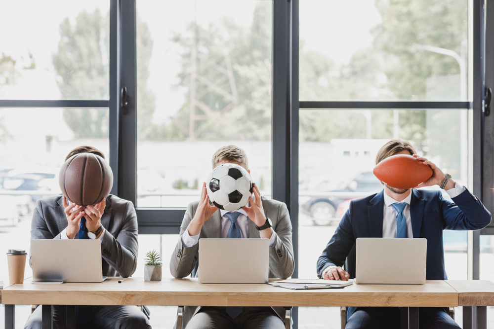 Men building sports betting models hold a basketball, soccer ball and football over their faces