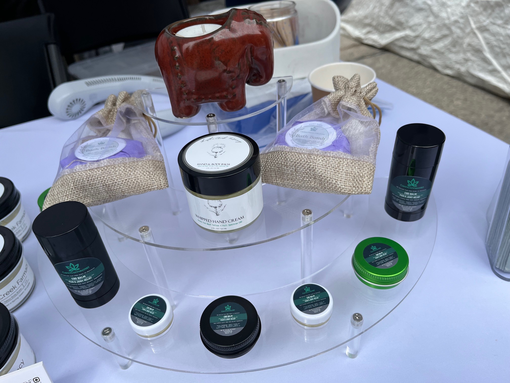 Clear acrylic display stand with various CBD balms, whipped hand cream, two bath bombs in burlap bags, and a small red elephant figurine on top.