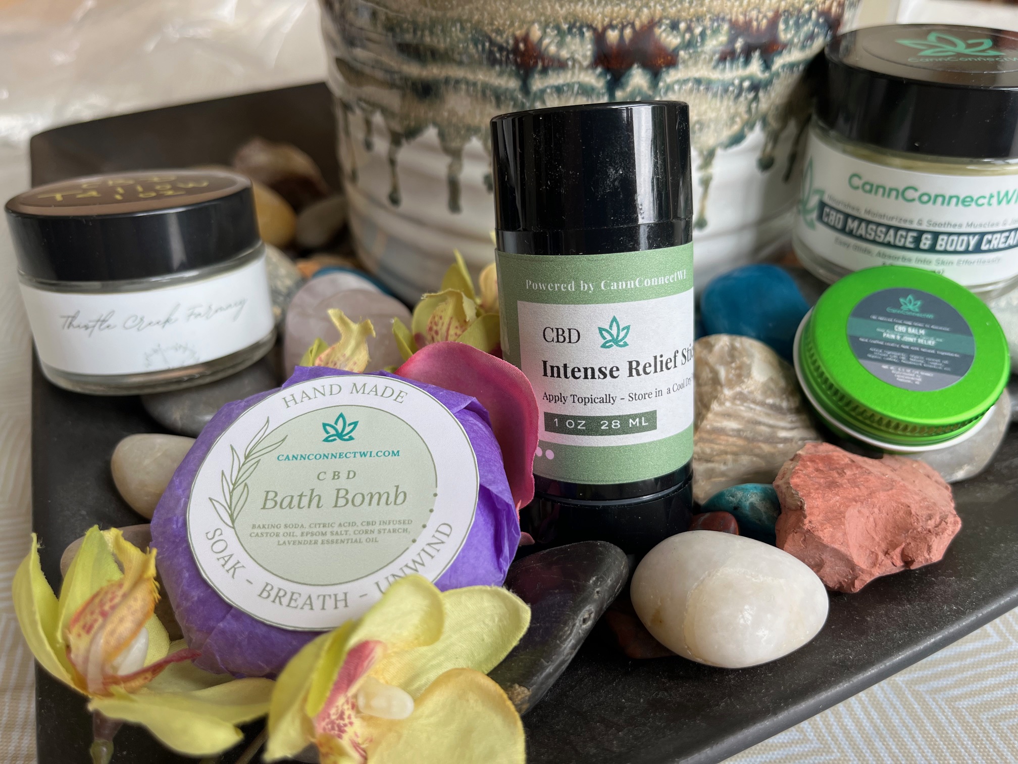 Display of CBD wellness products including a bath bomb, intense relief stick, massage and body cream, and CBD balm surrounded by stones and flowers.