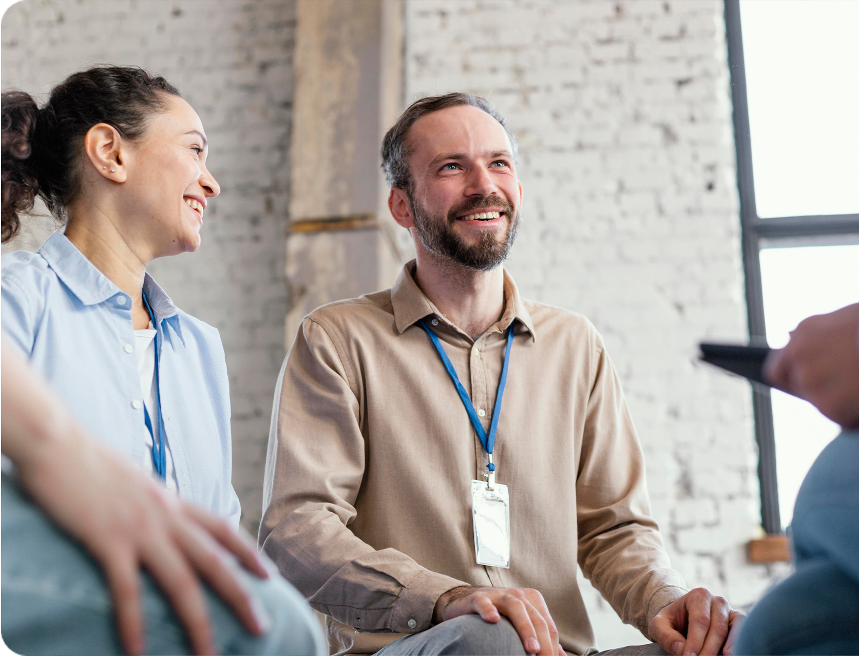 Two people with ID badges smiling and talking indoors against a brick wall background.