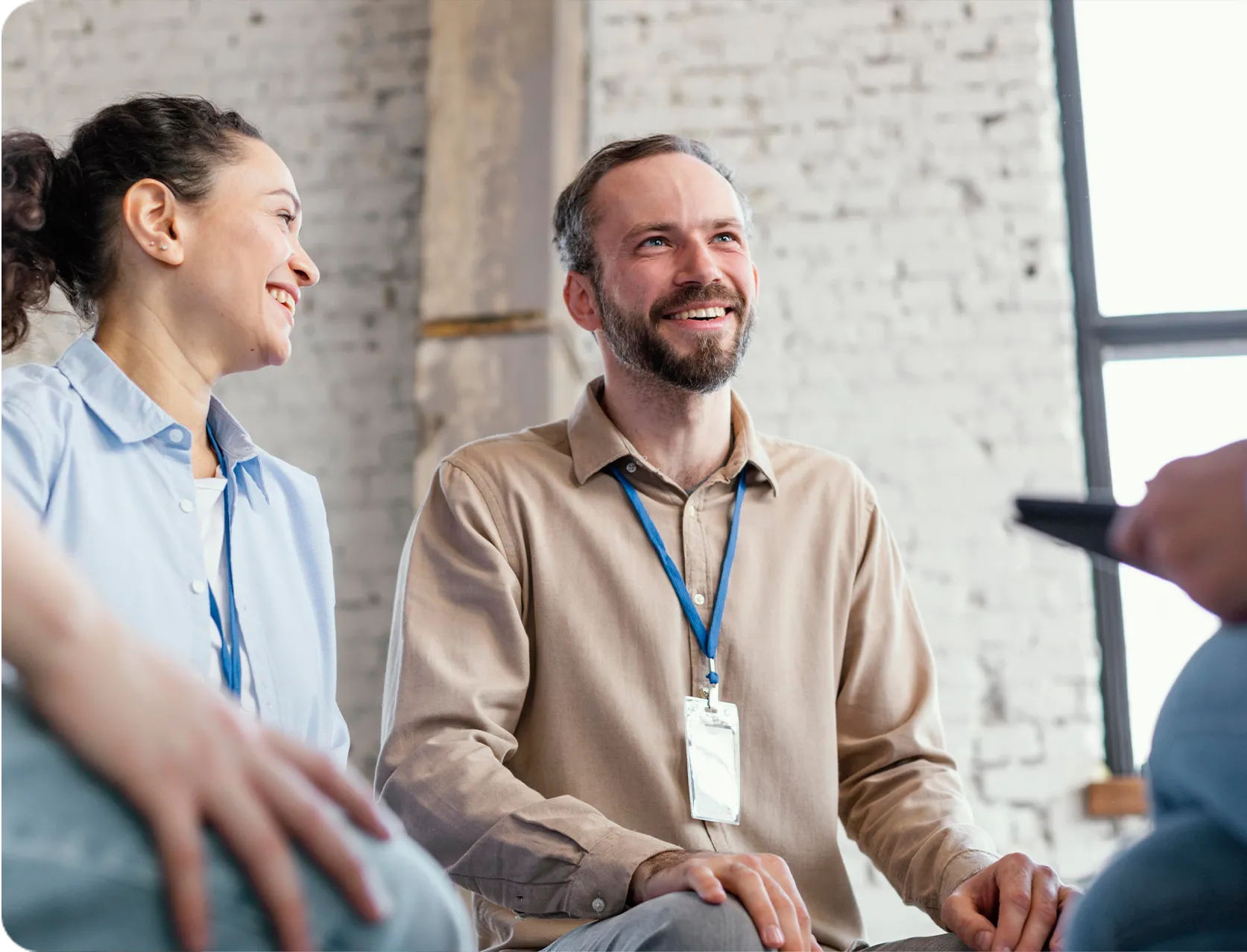 Two people with ID badges smiling and talking indoors against a brick wall background.