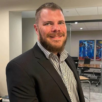 Smiling Dr. Heller wearing a black suit jacket and striped shirt in a modern office.