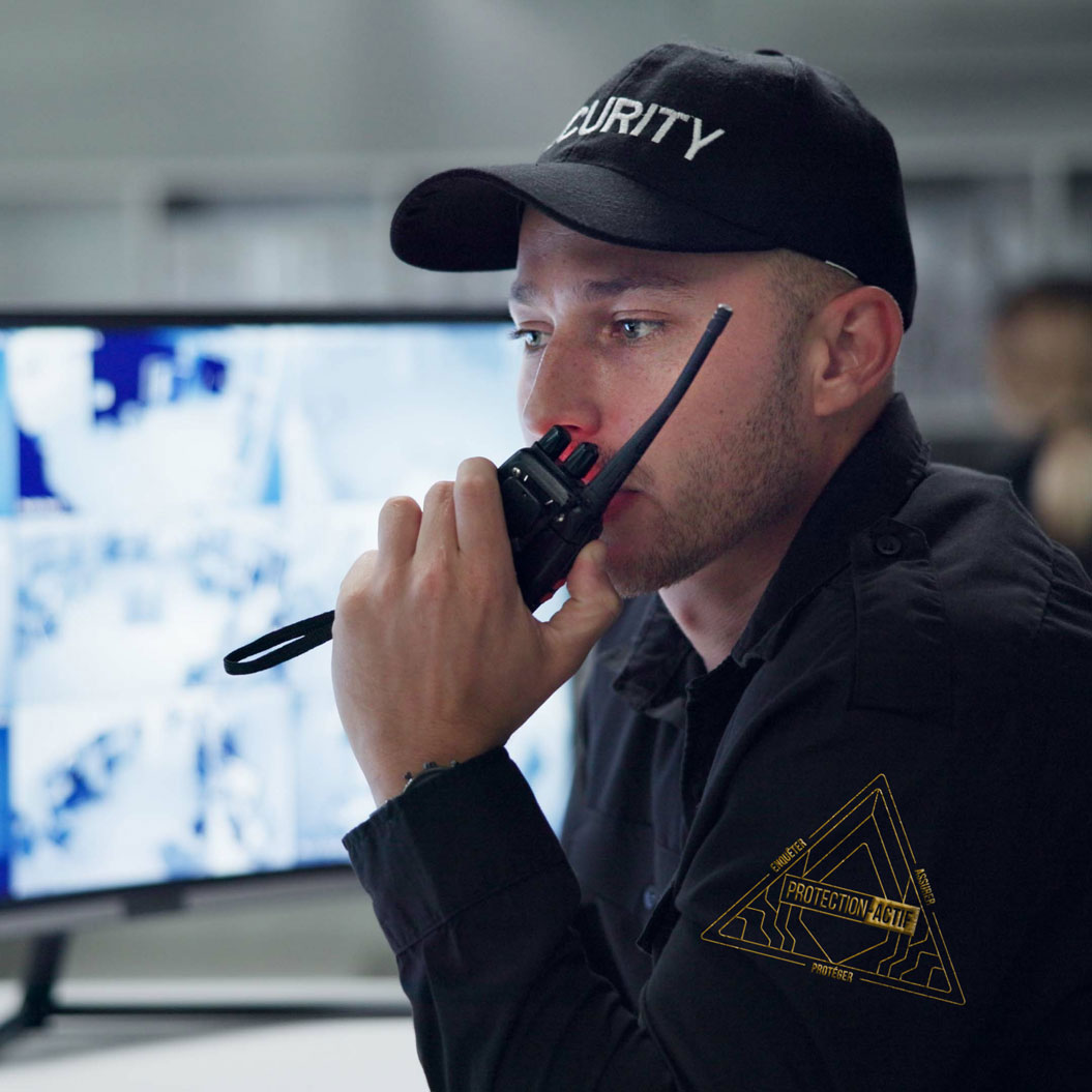 Agent de sécurité utilisant une radio pour communiquer dans une salle de surveillance avec des écrans CCTV en arrière-plan.