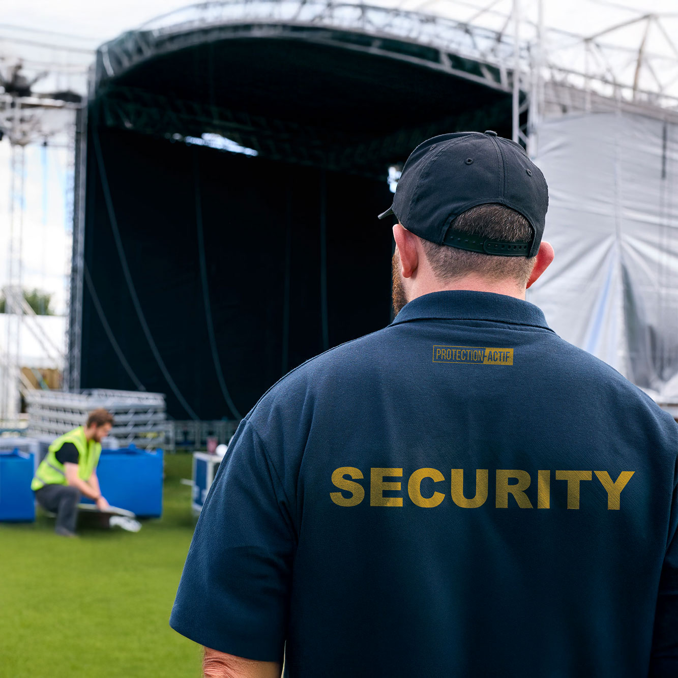 Rear view of a security guard in navy shirt and cap standing outdoors near an empty stage with another person working in the background.