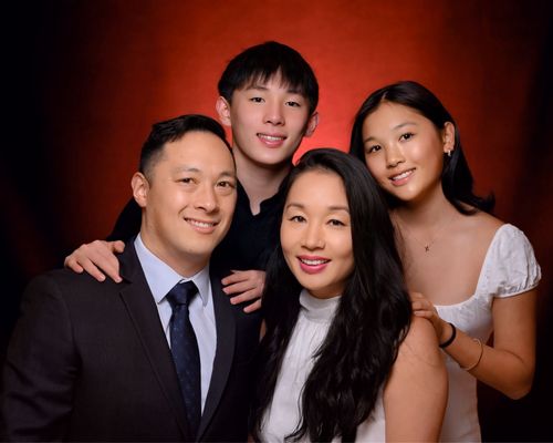 Portrait of a smiling family of four with a red gradient background, father in suit and tie, mother in white top, son and daughter standing behind them.