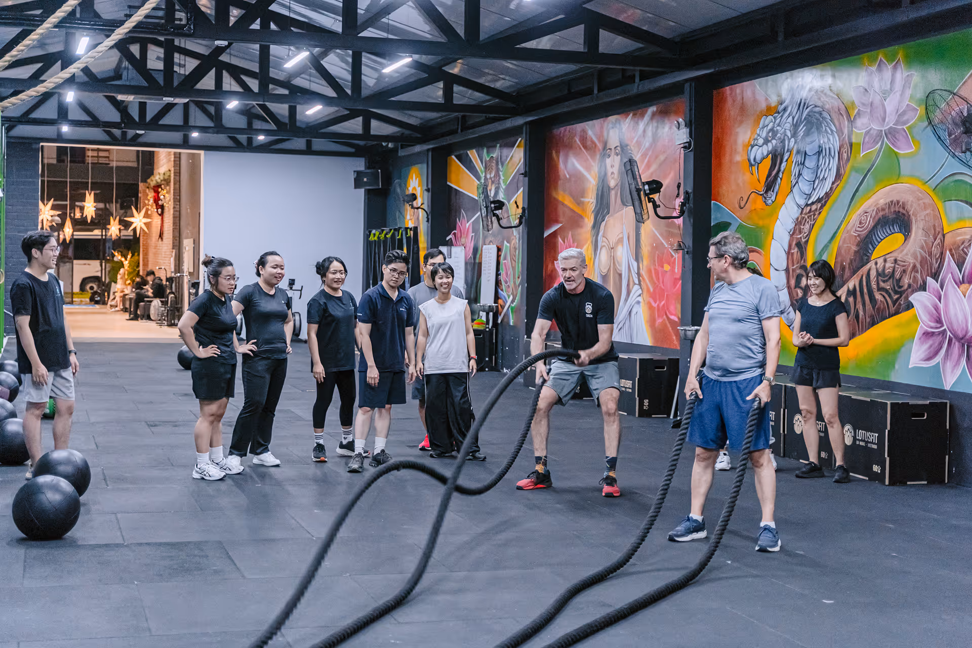 Two men in gym using battle ropes while others watch and mural art decorates the wall.