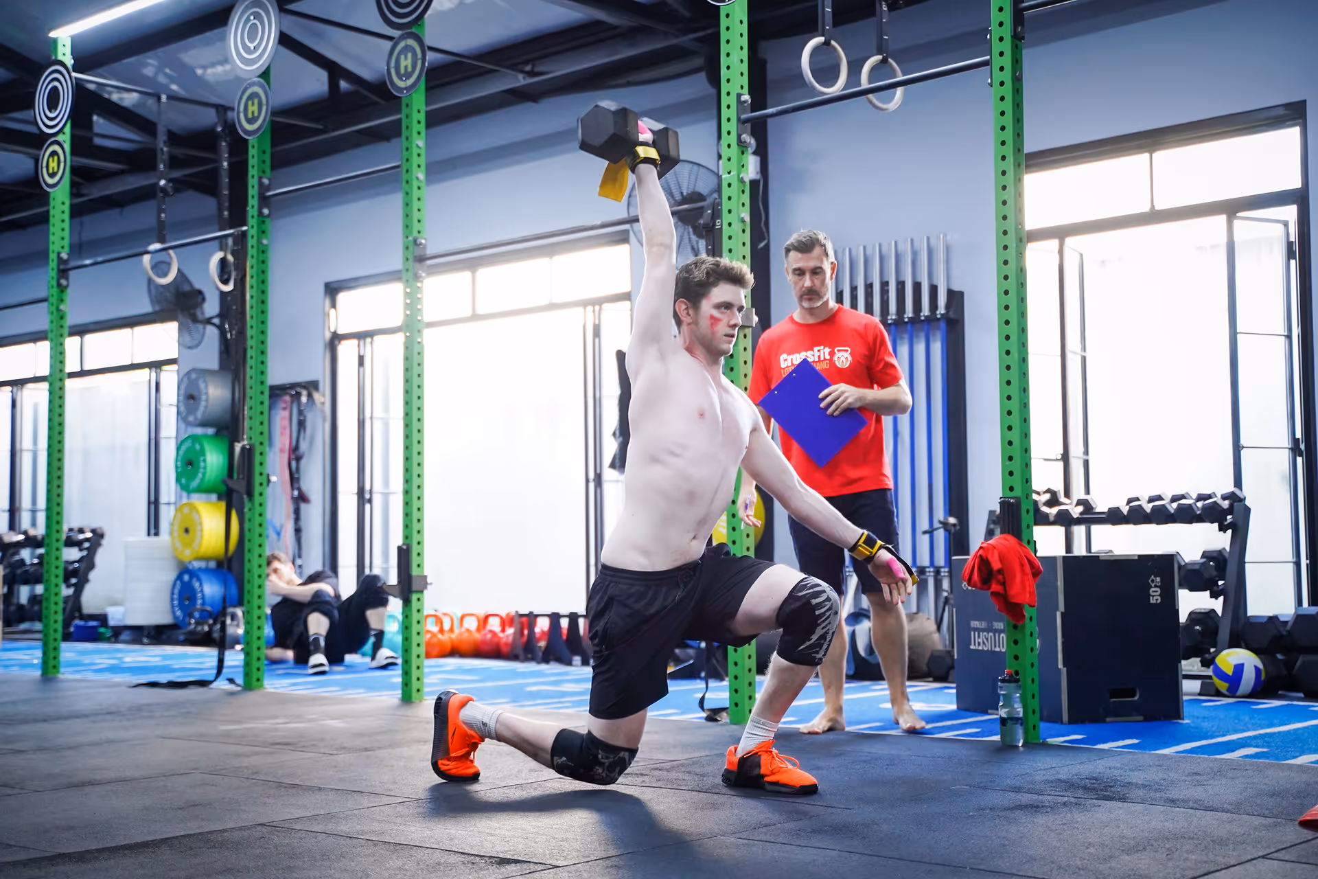 Shirtless man in black shorts and orange shoes performing a kneeling dumbbell press in a gym while a trainer in a red CrossFit shirt observes with a clipboard.