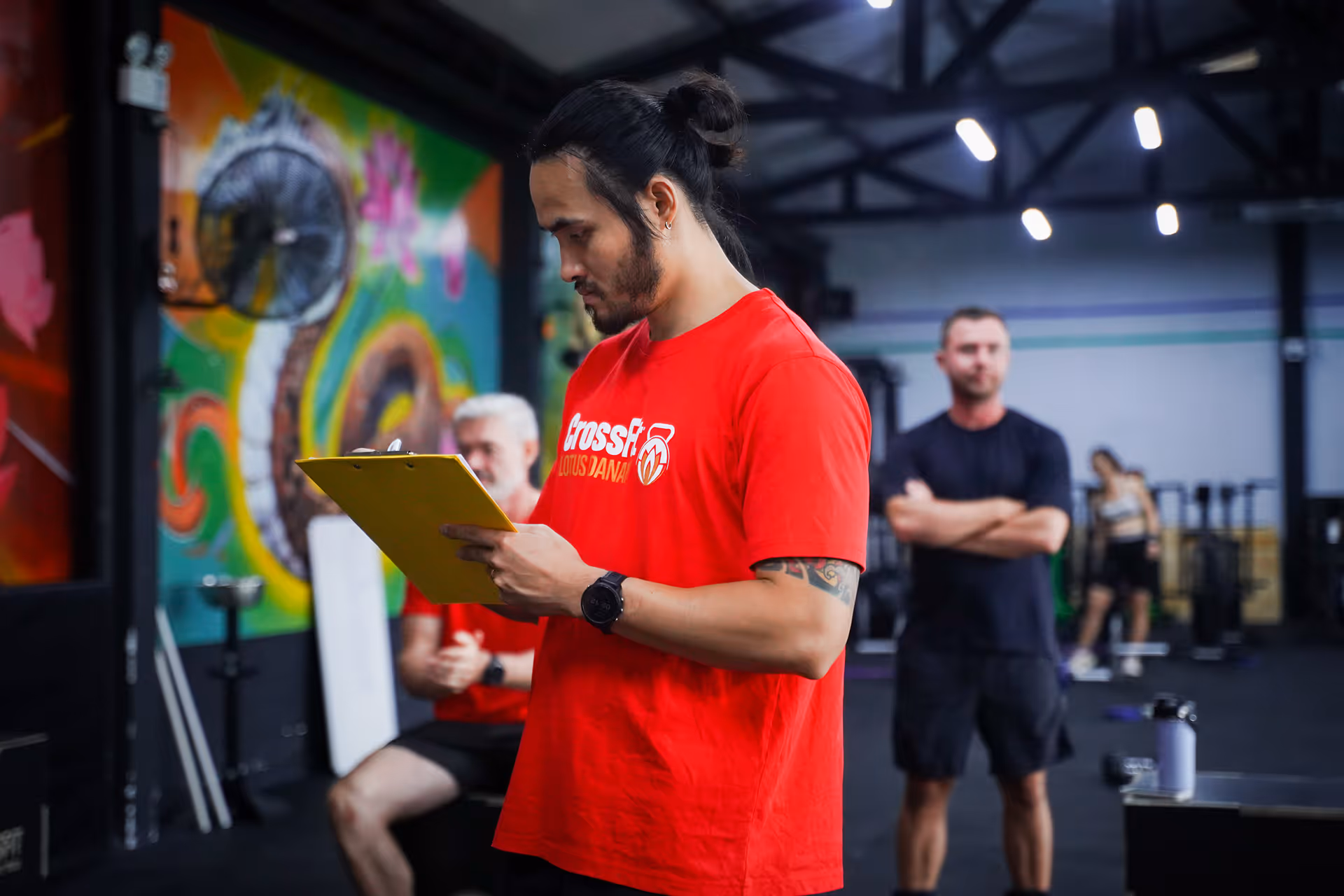 Man in red CrossFit shirt holding yellow clipboard in gym with others in background.