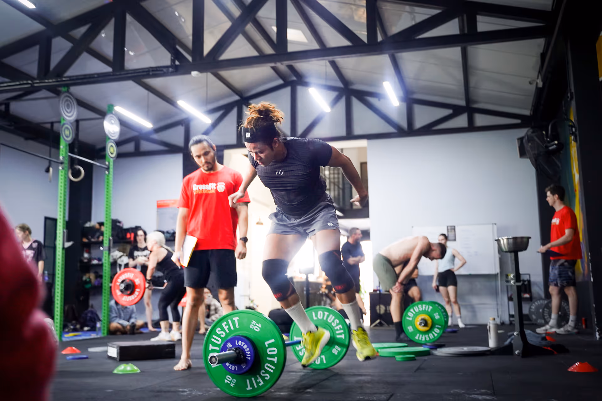 Person jumping over barbell plates in a CrossFit gym with coach observing and other people training in background.