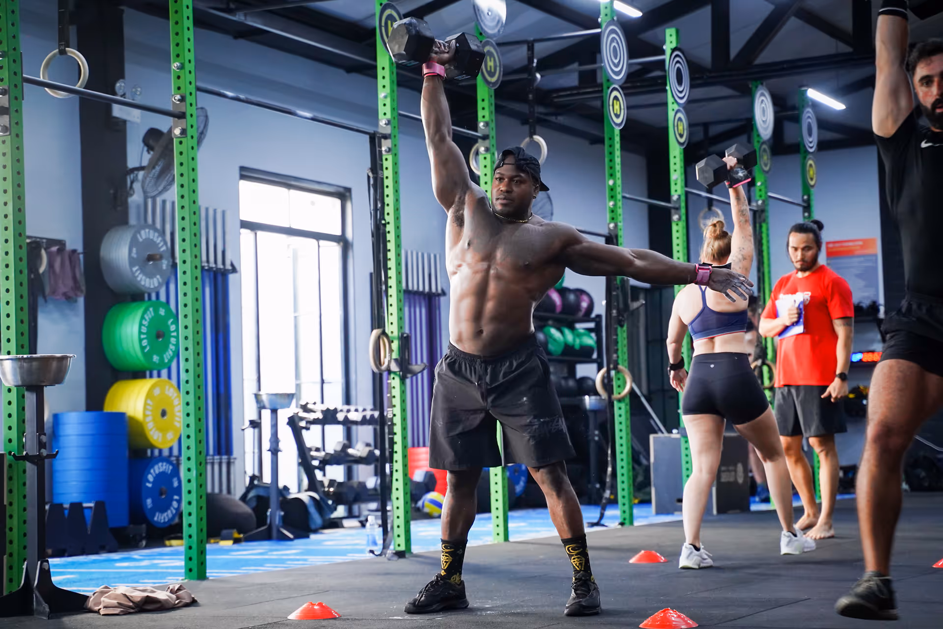 Muscular man lifting a dumbbell overhead during a workout in a gym with other people exercising nearby.
