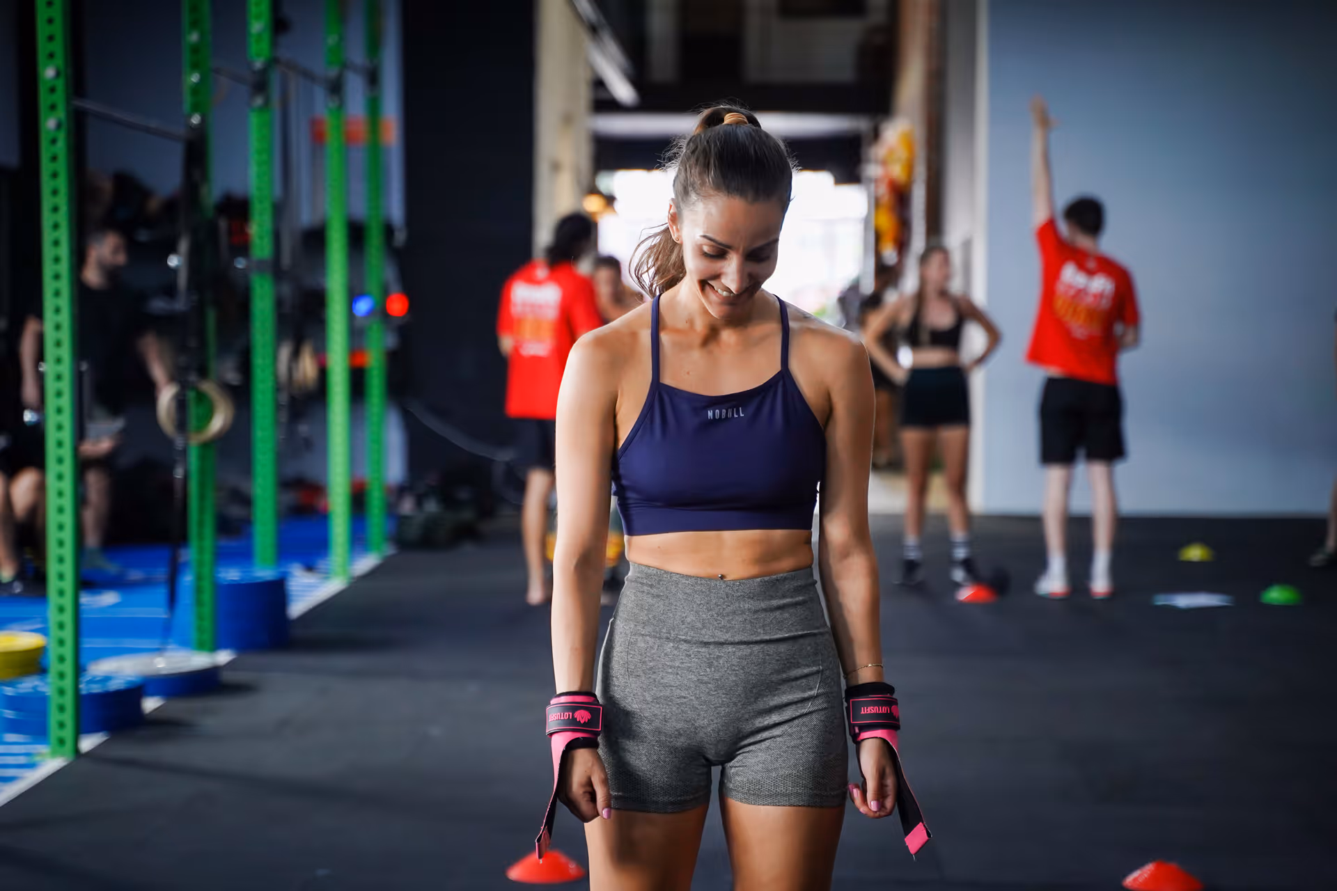 Smiling woman in workout attire with wrist wraps standing in a gym with others exercising in the background.