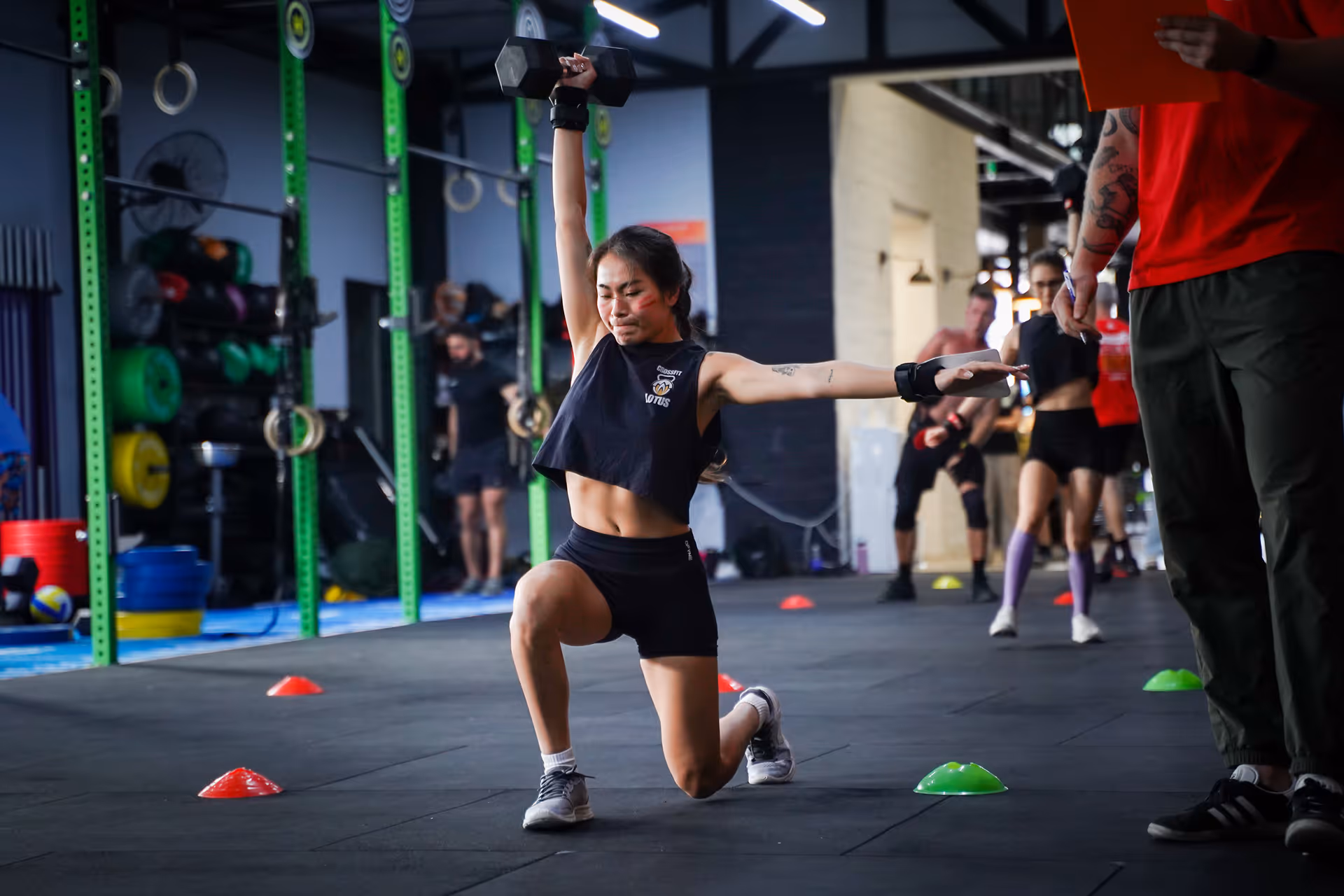 Woman performing a one-arm dumbbell overhead lunge in a gym with others and a coach holding a clipboard nearby.