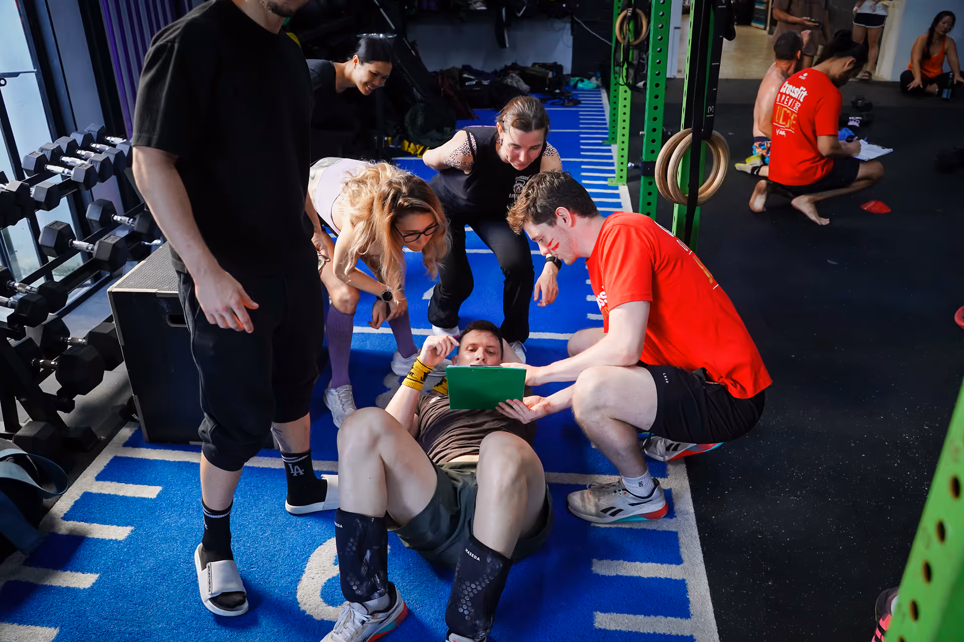 Group of people in a gym surrounding a man lying on a blue mat as they look at a green clipboard.