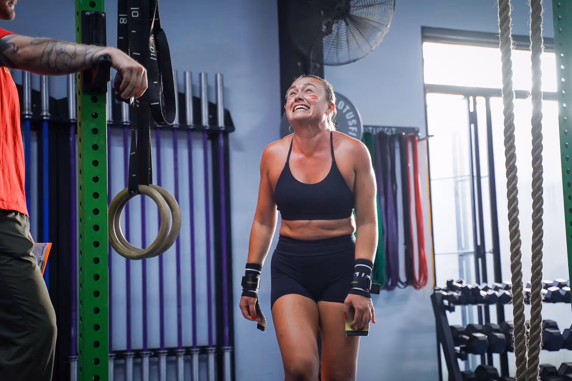 Smiling woman in black workout attire standing in a gym near gymnastic rings and weightlifting equipment.