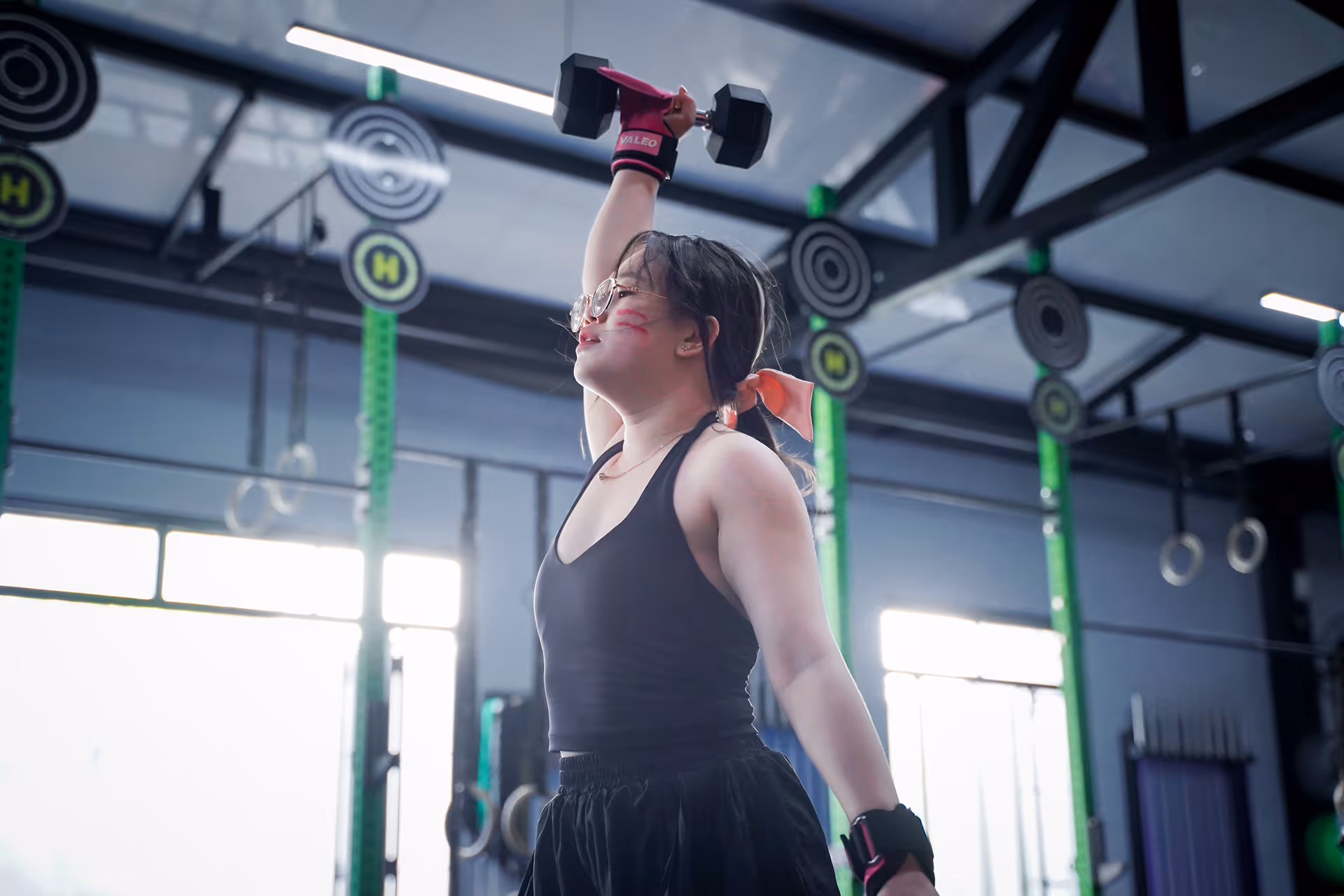 Woman lifting a dumbbell overhead in a gym, wearing black workout clothes and glasses.