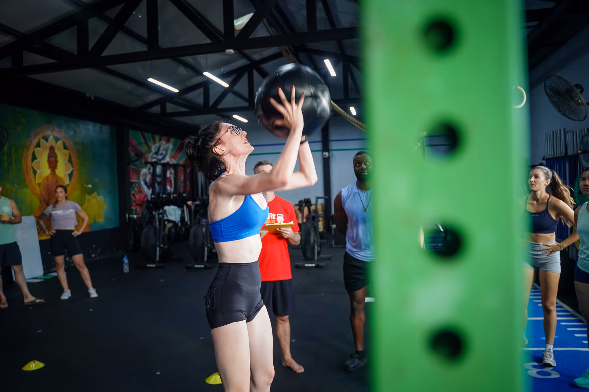 Woman in blue sports bra and black shorts lifting a medicine ball in a gym with people watching.