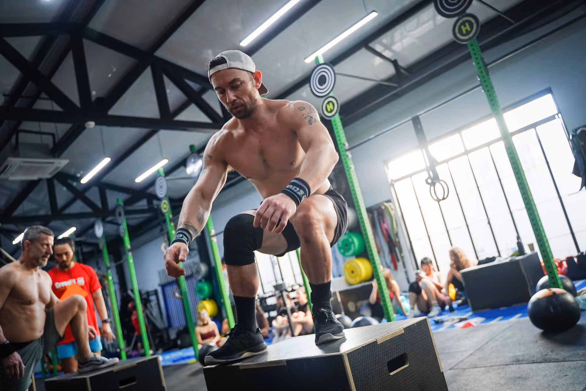 Shirtless man wearing a backwards cap performing a box jump exercise in a gym with others around.