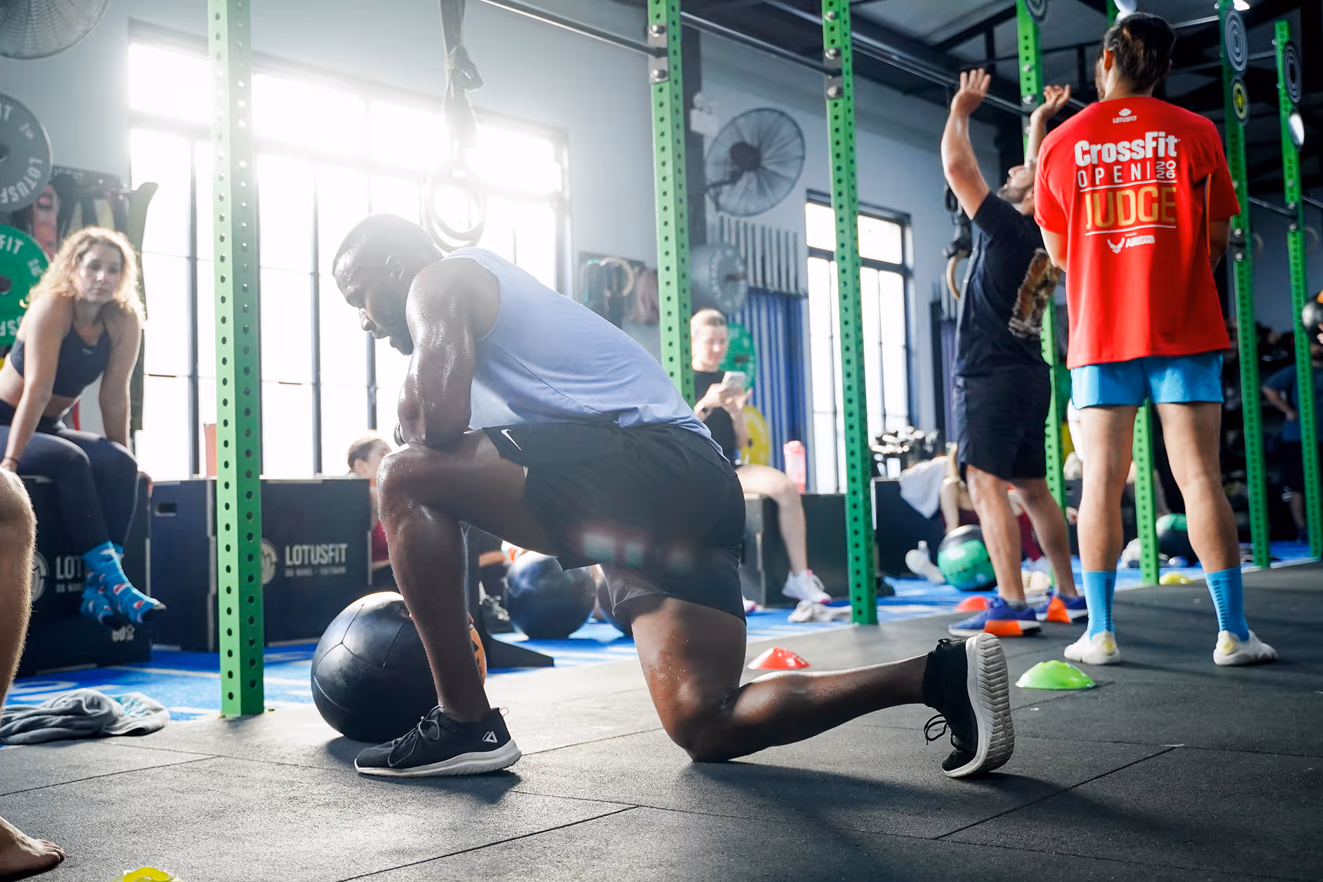 Man in white tank top and black shorts stretching in a gym with other people exercising in the background.
