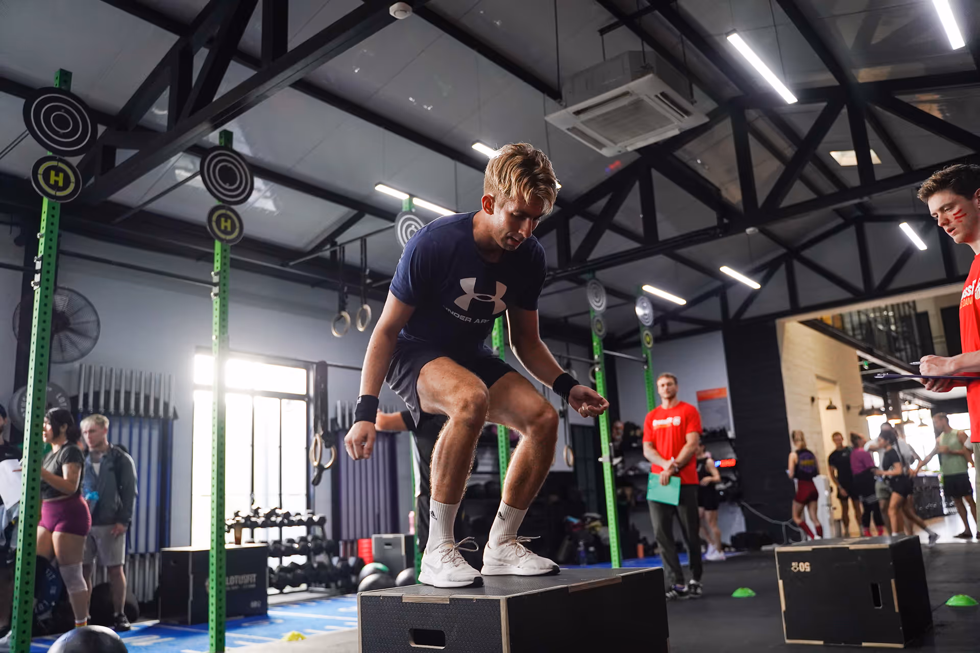 Young man performing a box jump exercise in a busy gym with people and fitness equipment in the background.