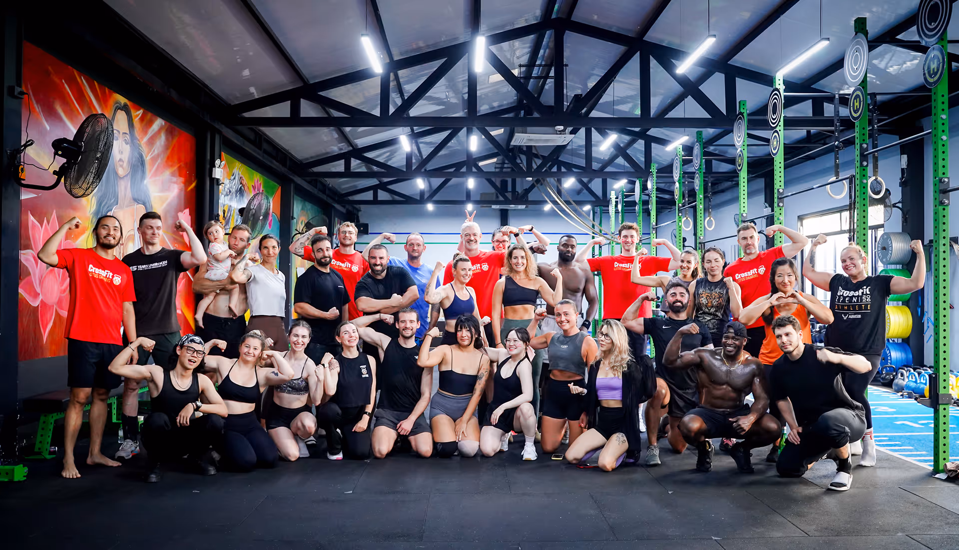 Large diverse group of people posing and flexing muscles inside a bright CrossFit gym.