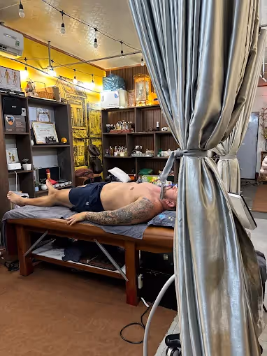 Tattooed man lying on a treatment table in a cozy room with shelves and string lights.