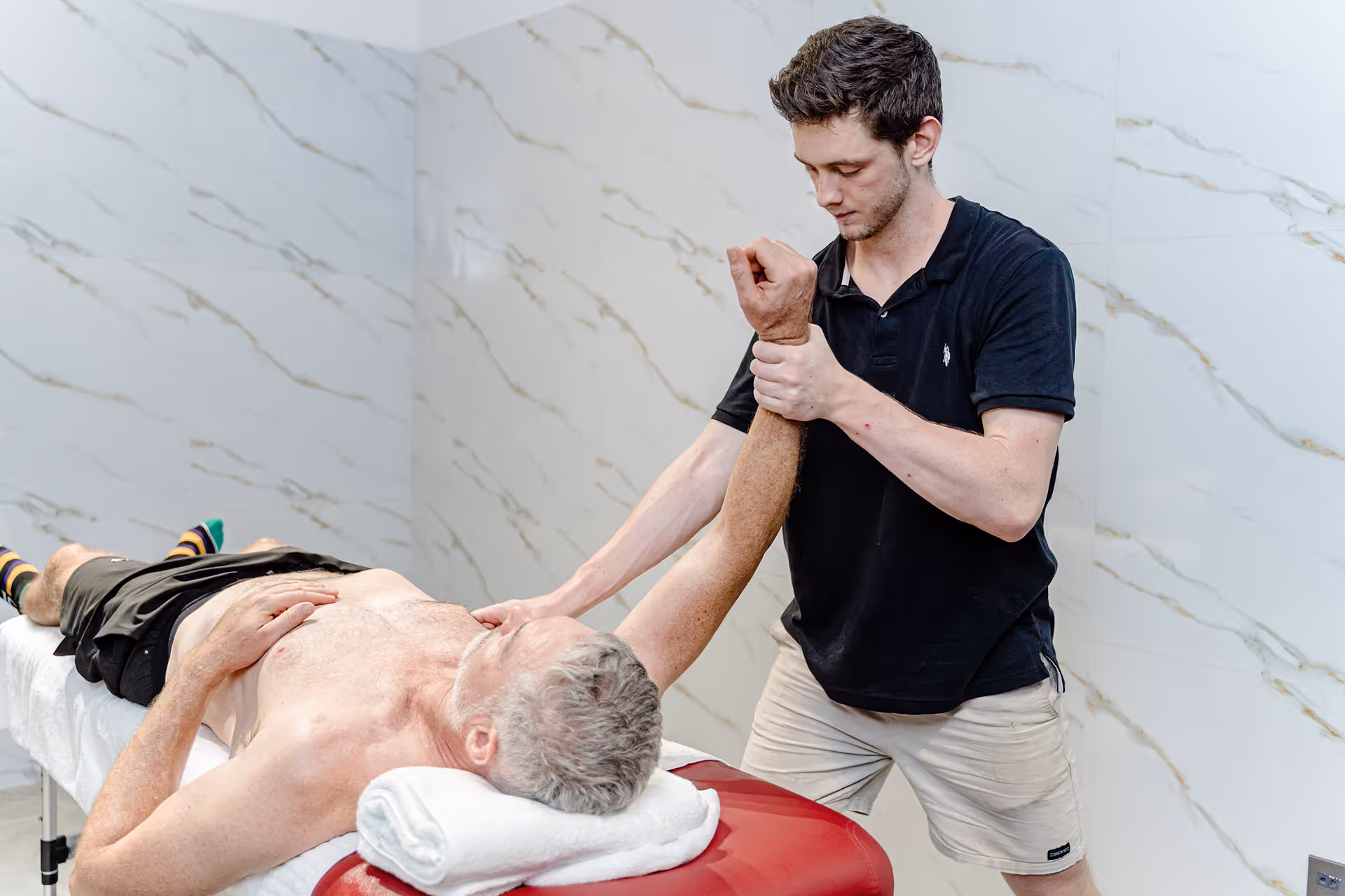 Therapist in black shirt stretches older man's arm on massage table in a room with marble walls.