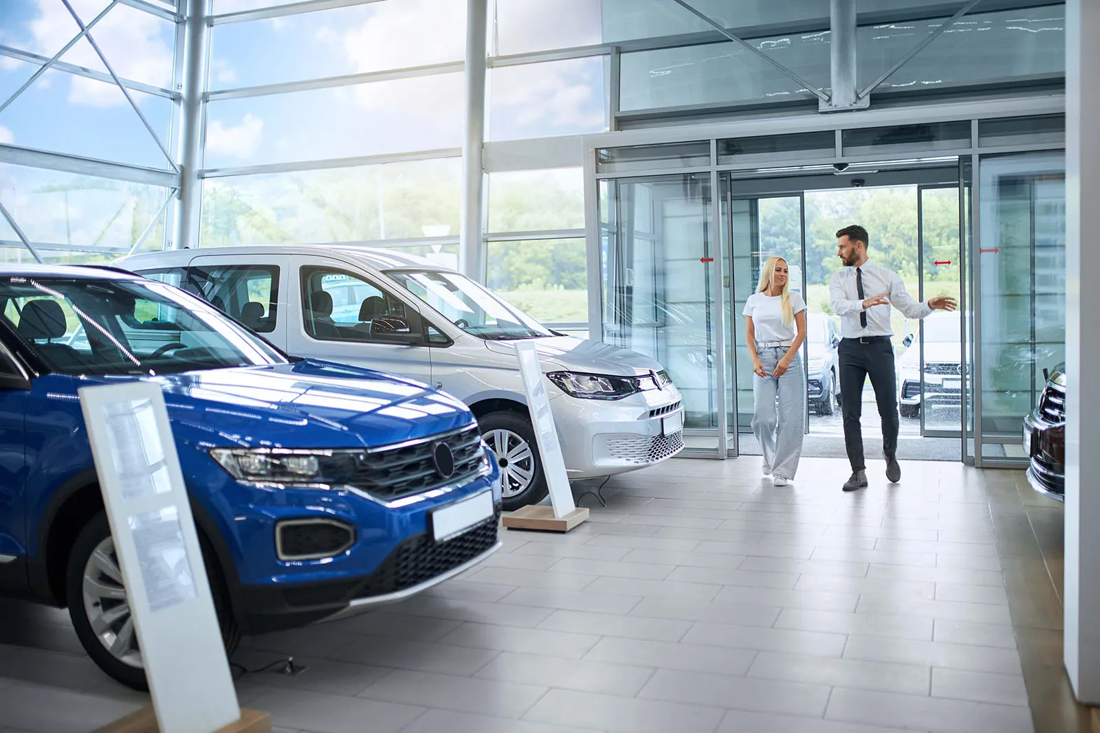 A car salesperson walking with a female customer inside a brightly lit dealership showroom, with several cars, including a blue SUV, parked on display.