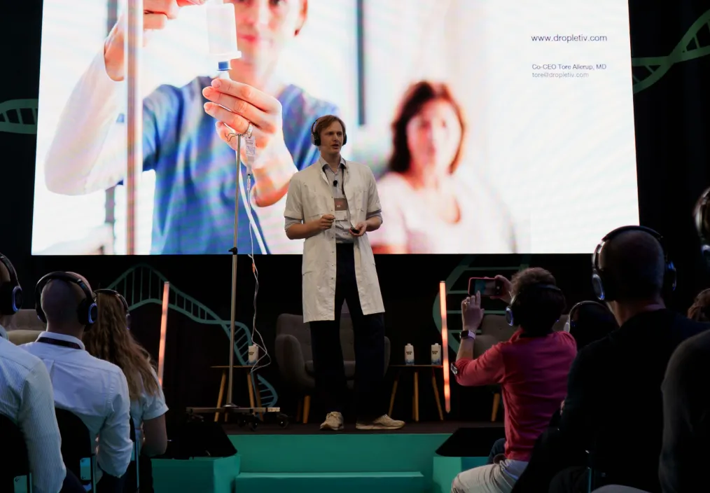 A speaker in a white lab coat presenting on stage in front of a large screen showing a medical professional handling an IV, with an audience seated and some recording the talk.