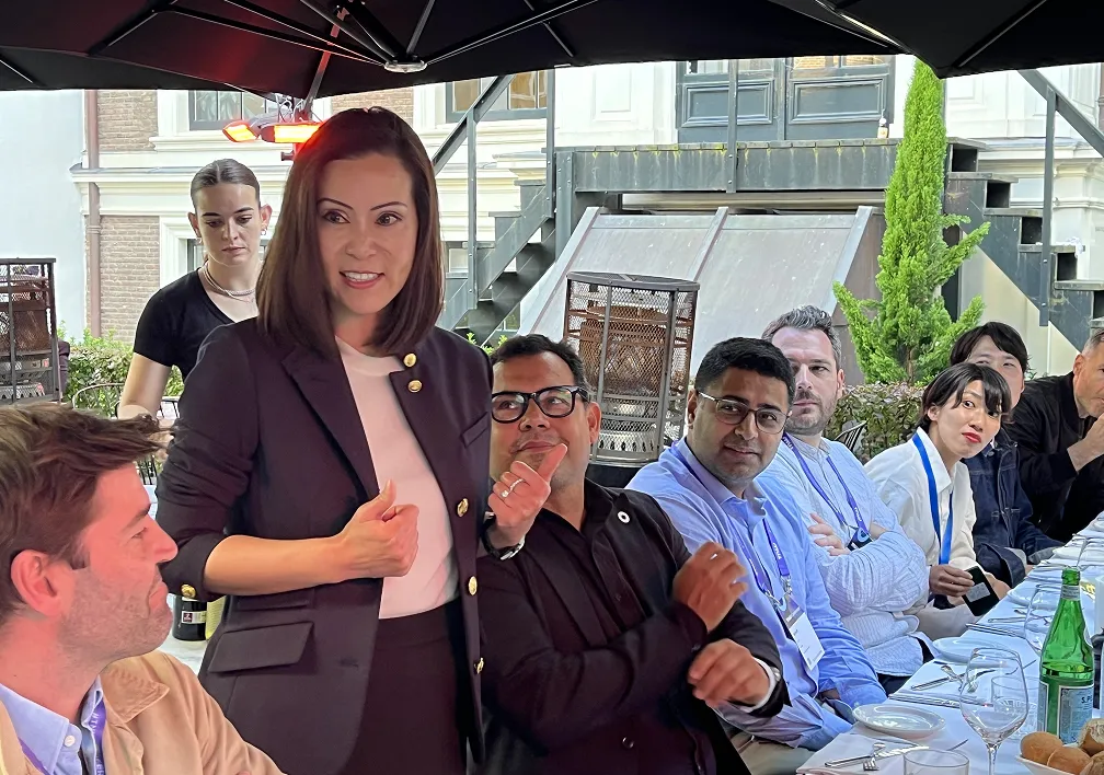 A woman standing and speaking at an outdoor dinner table with a group of seated attendees listening attentively.