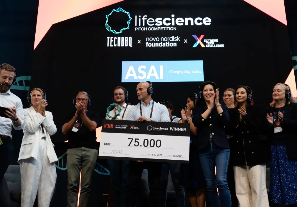 A group of people on stage celebrating the winner of the Life Science Pitch Competition, with the central person holding an oversized check for 75,000 DKK, under event branding for TechBBQ, Novo Nordisk Foundation, ASAI, and Extreme Tech Challenge.