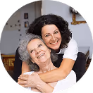 Glückliche ältere Frau und jüngere Frau umarmen sich lächelnd zu Hause im Wohnzimmer.