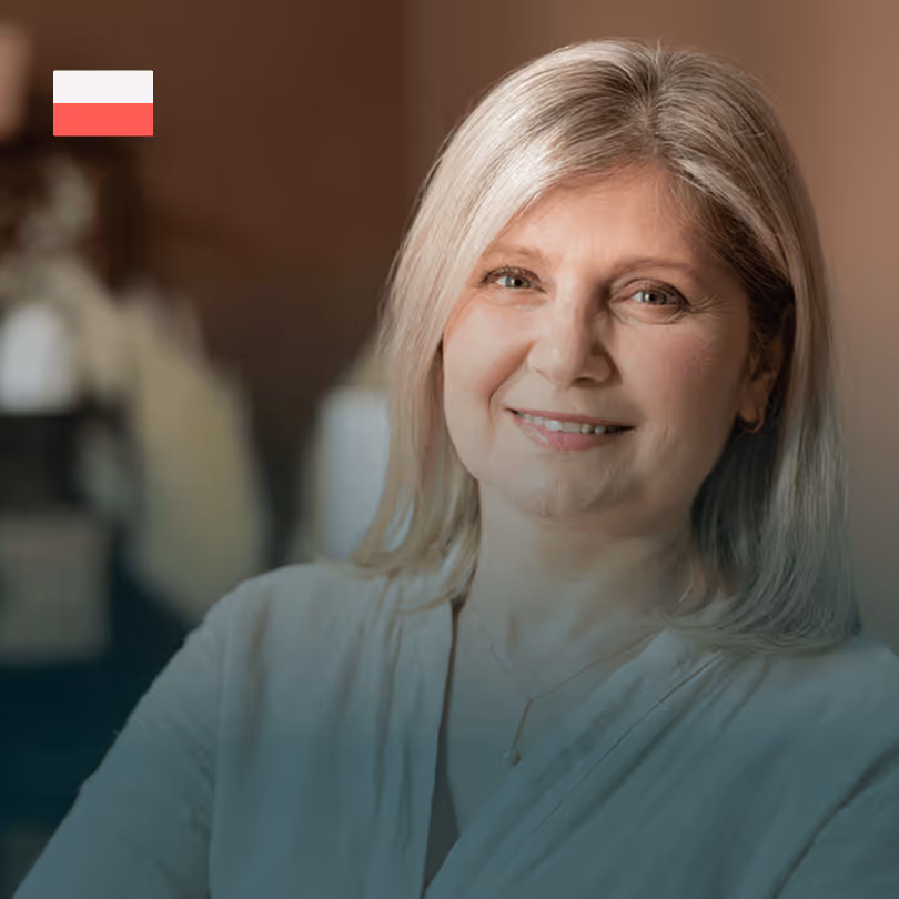 Ältere Frau mit graublondem Haar lächelt, polnische Flagge in der Ecke, warme Raumbeleuchtung.