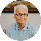 Älterer Mann mit Brille und weißem Haar lächelt, trägt ein hellblaues Polohemd, sitzt drinnen vor dezentem Hintergrund.