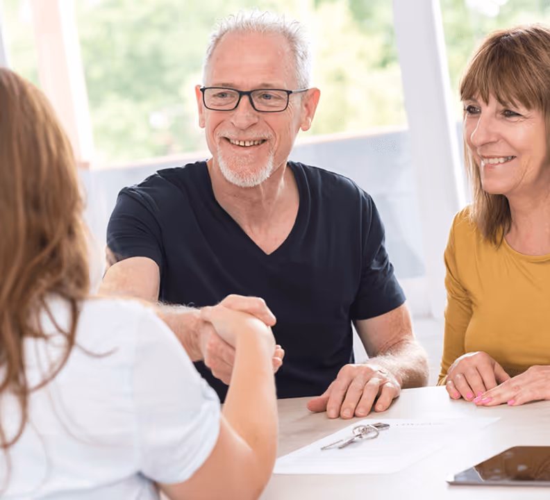 Älteres Paar lächelt und gibt junger Frau die Hand, während sie am Tisch mit Dokumenten und Schlüssel sitzen.