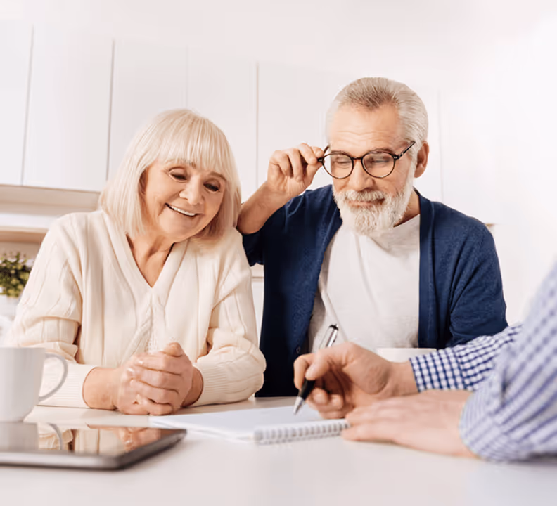 Älteres Paar sitzt lächelnd am Tisch, hört einer Person zu, die auf ein Notizbuch zeigt, Tablet und Tasse daneben.