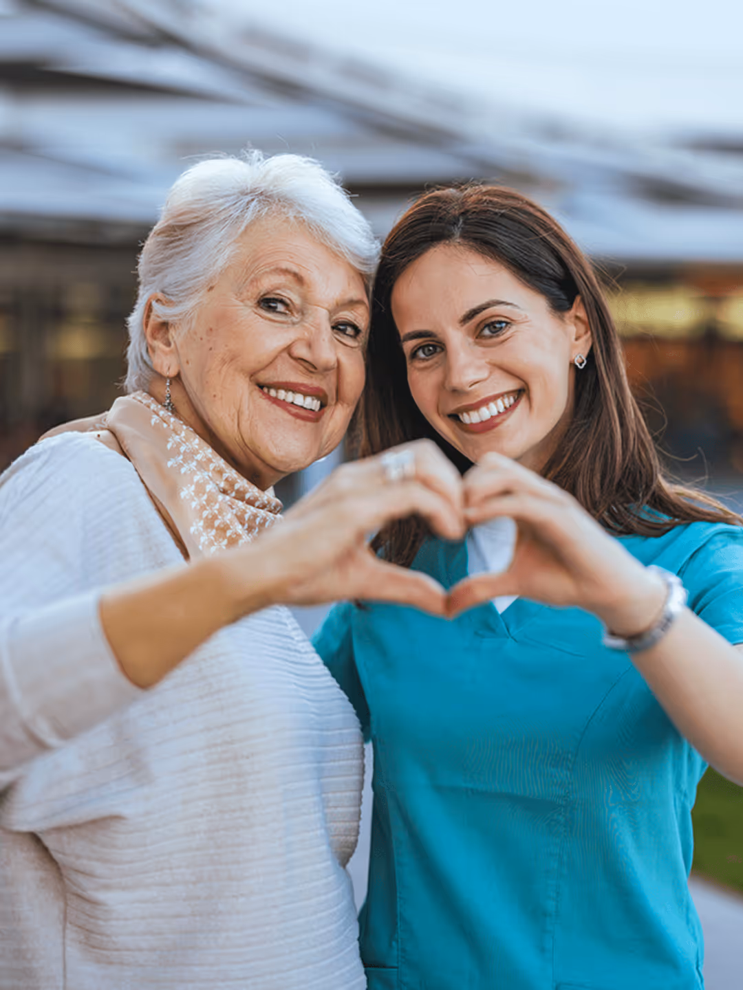 Ältere Frau und jüngere Frau in blauer Uniform formen gemeinsam ein Herz mit ihren Händen und lächeln in die Kamera.