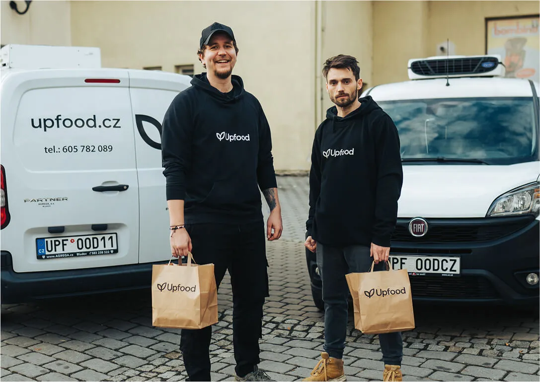 Dva muži v černých mikinách Upfood drží papírové tašky Upfood, stojí mezi dvěma bílými dodávkami s logem upfood.cz.