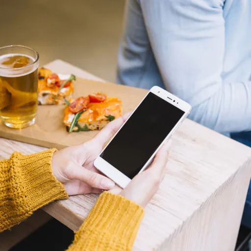 Person holding a white smartphone with a blank screen at a table with two slices of pizza and a glass of beer.