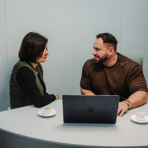 Two people sitting at a table with a laptop and two cups of coffee, engaged in conversation.
