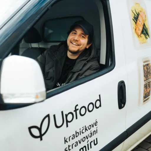 Smiling man wearing a black cap sitting in a white Upfood delivery van with Czech text and food images on the side.